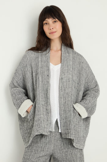A woman with long brown hair wears the Crinkle Double Face Linen Jacket in gray over a white top and matching pants, standing against a plain light background.