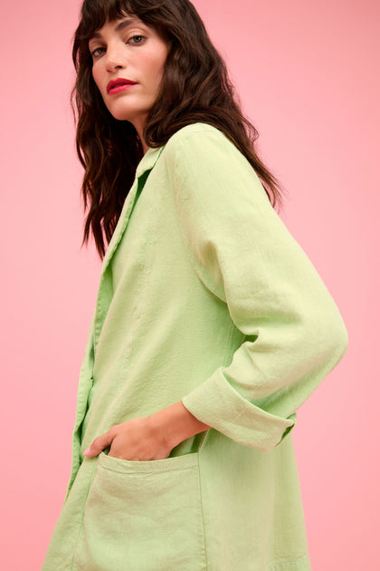 A woman with long dark hair wears the Twisted Linen Relaxed Blazer with an oversized fit, standing against a pink background and looking at the camera with one hand in her pocket.