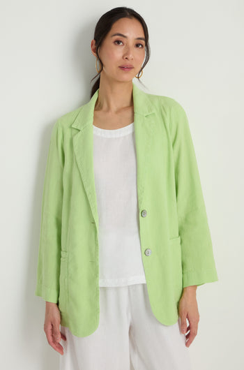 Model wears the Twisted Linen Relaxed Blazer in light green over a white top and white pants, standing against a plain light-colored background.