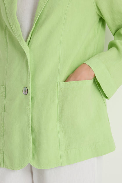 A person wears the Twisted Linen Relaxed Blazer in light green, styled with one hand in the front pocket and paired with white pants.