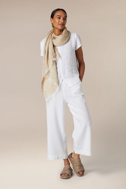 A woman stands in front of a neutral background wearing white casual summer clothing, including a sleeveless top, Texture Linen Wide Trouser with an elasticated waist, and beige sandals, complemented by a light scarf