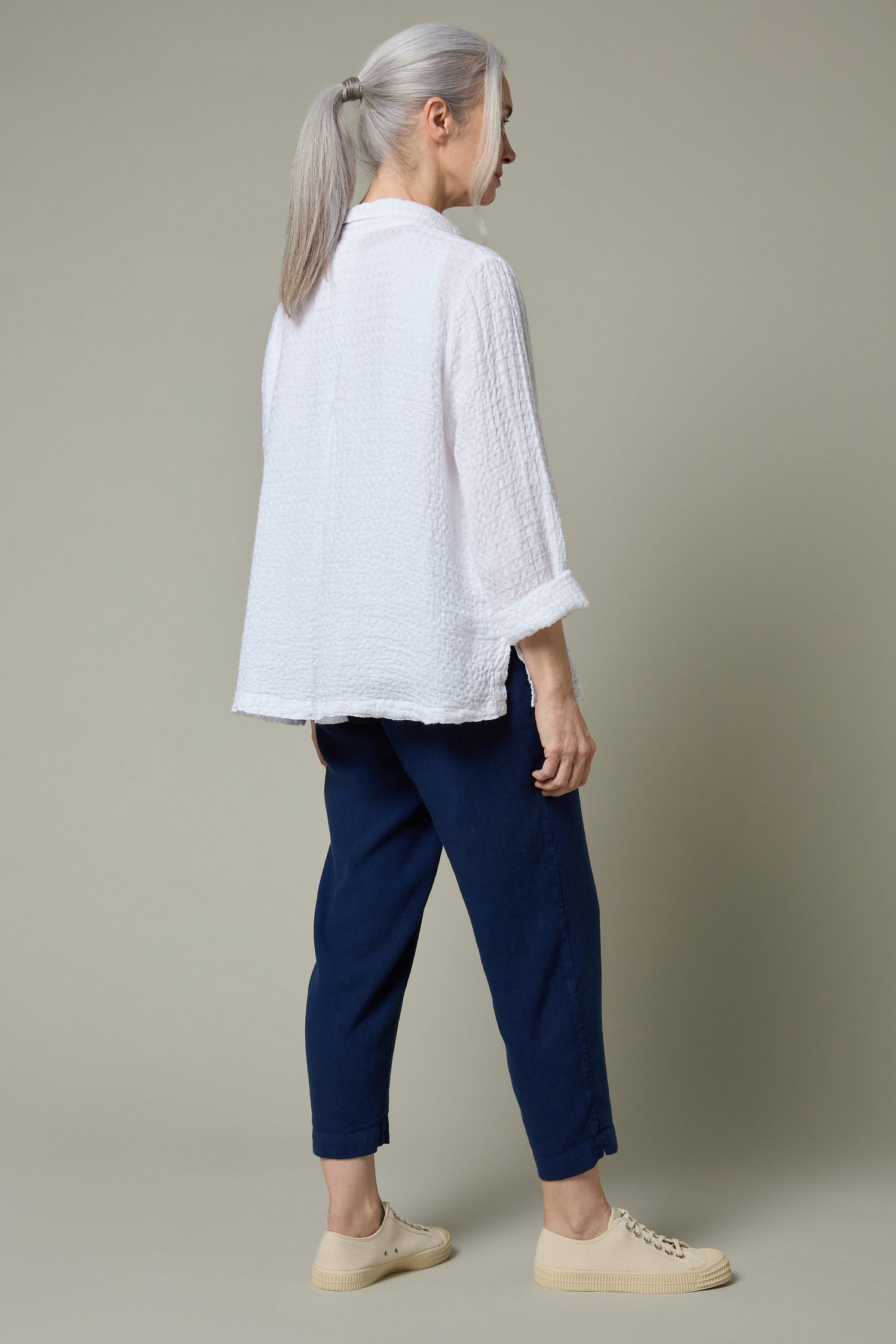An older adult with gray hair in a ponytail wears a loose white linen top, Twisted Linen Navy Slim Trouser with an elasticated waistband, and beige sneakers, standing sideways against a plain background.