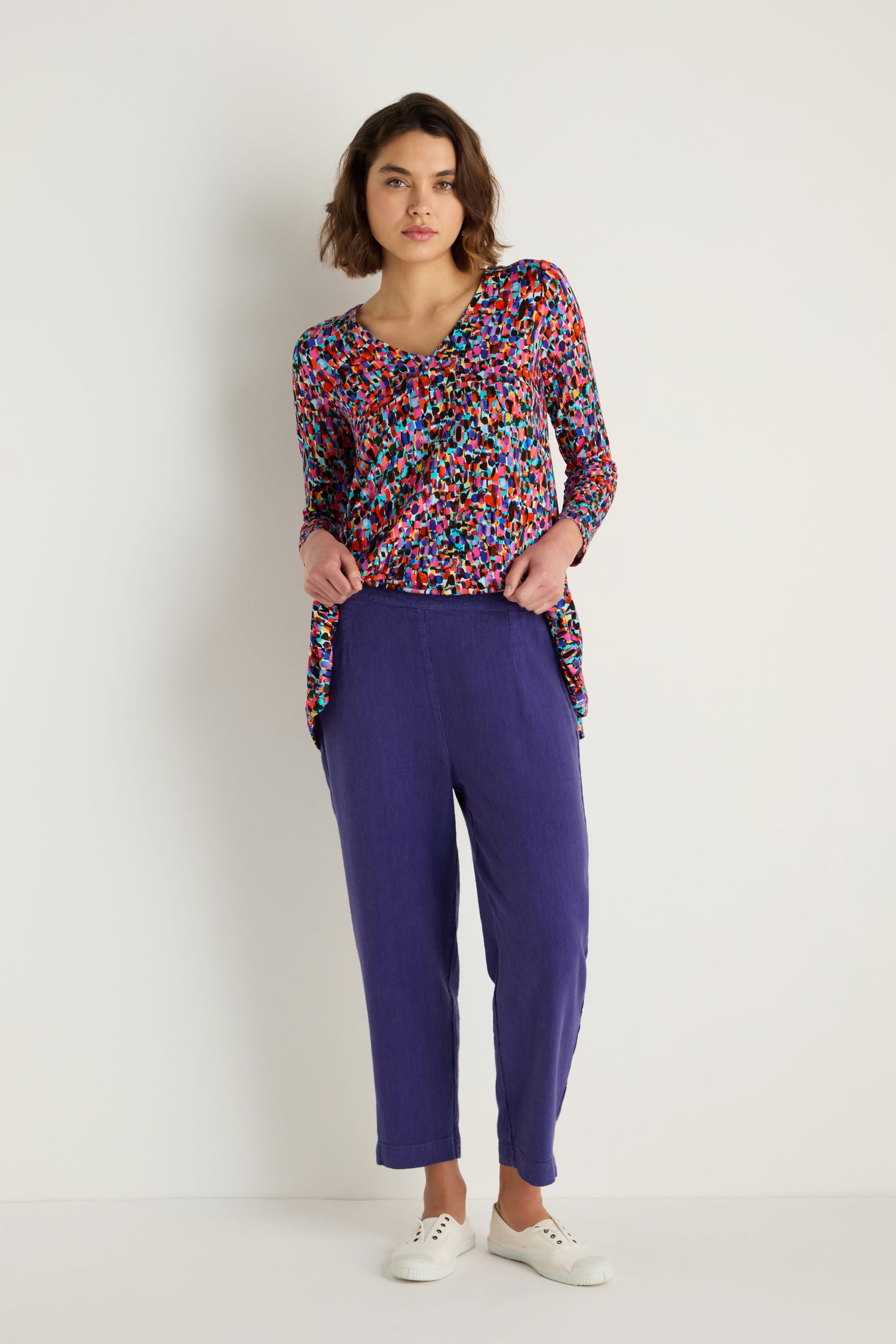 A woman stands against a white background wearing a colorful long-sleeve top, Twisted Linen Slim Trouser in purple, and white sneakers, with her hands in her pockets.