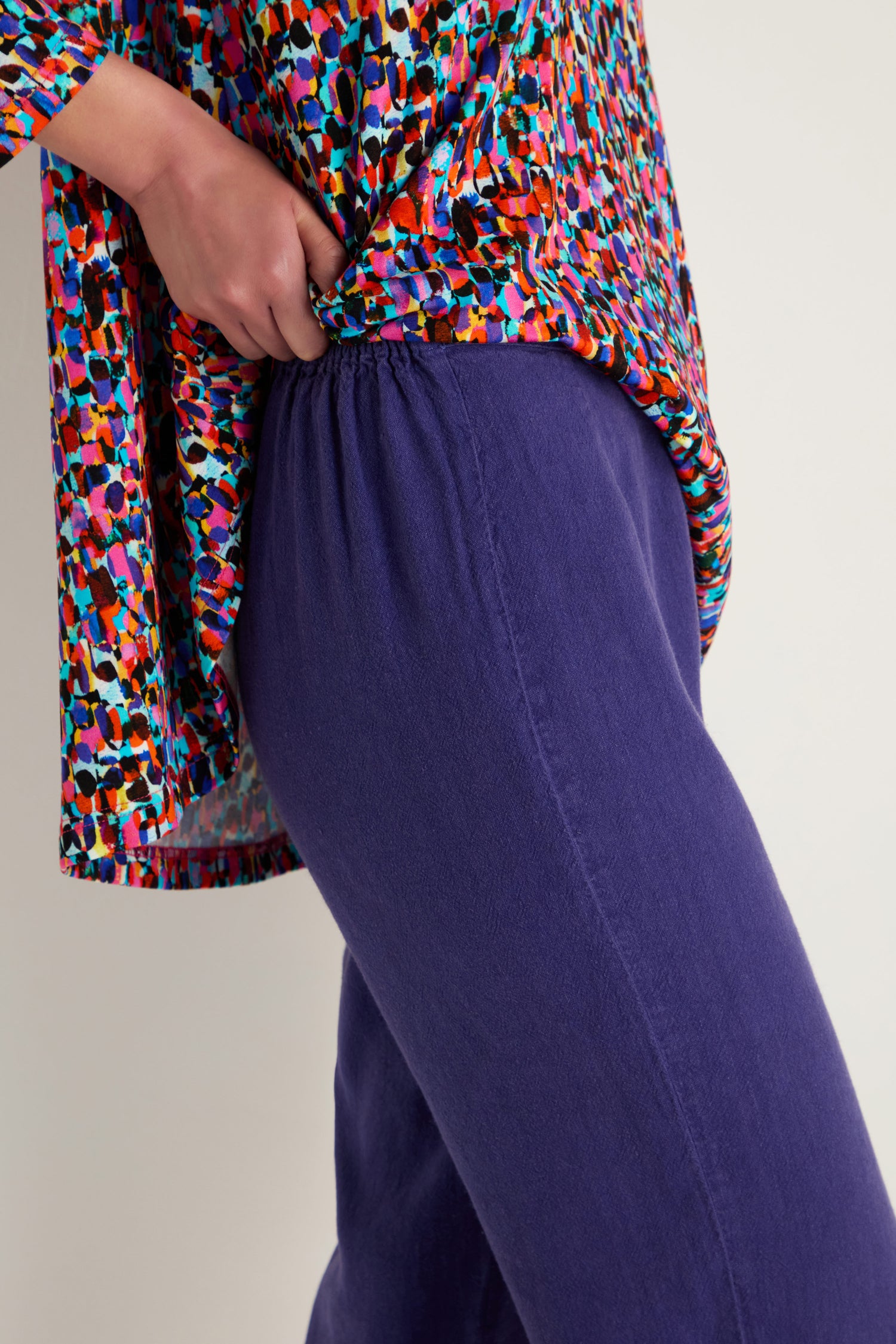 A person wearing the Twisted Linen Slim Trouser in purple and a colorful patterned top, adjusting the waistband with one hand.