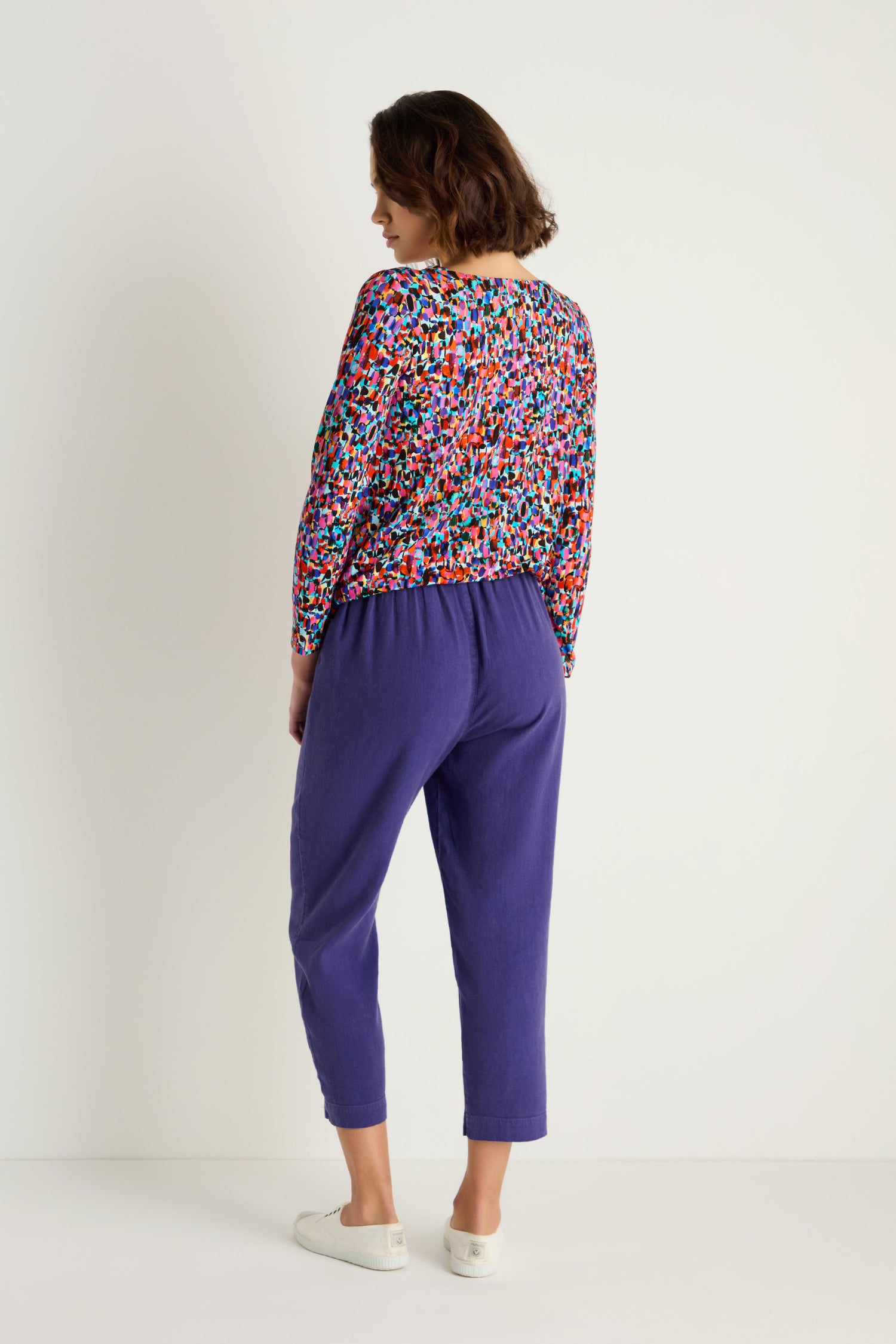 A woman faces away in a colorful long-sleeve top, paired with Twisted Linen Slim Trouser in purple and white sneakers, set against a plain white background.