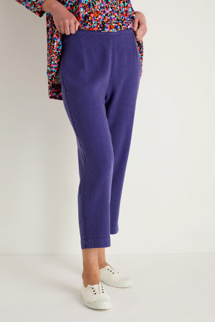 A person wearing Twisted Linen Slim Trousers in purple, a colorful patterned top, and white sneakers stands against a plain white background.