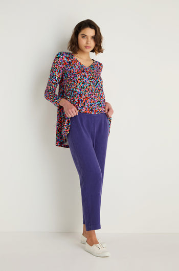 A woman stands against a white wall wearing a multicolored patterned long-sleeve top, Twisted Linen Slim Trouser in purple, and white sneakers.