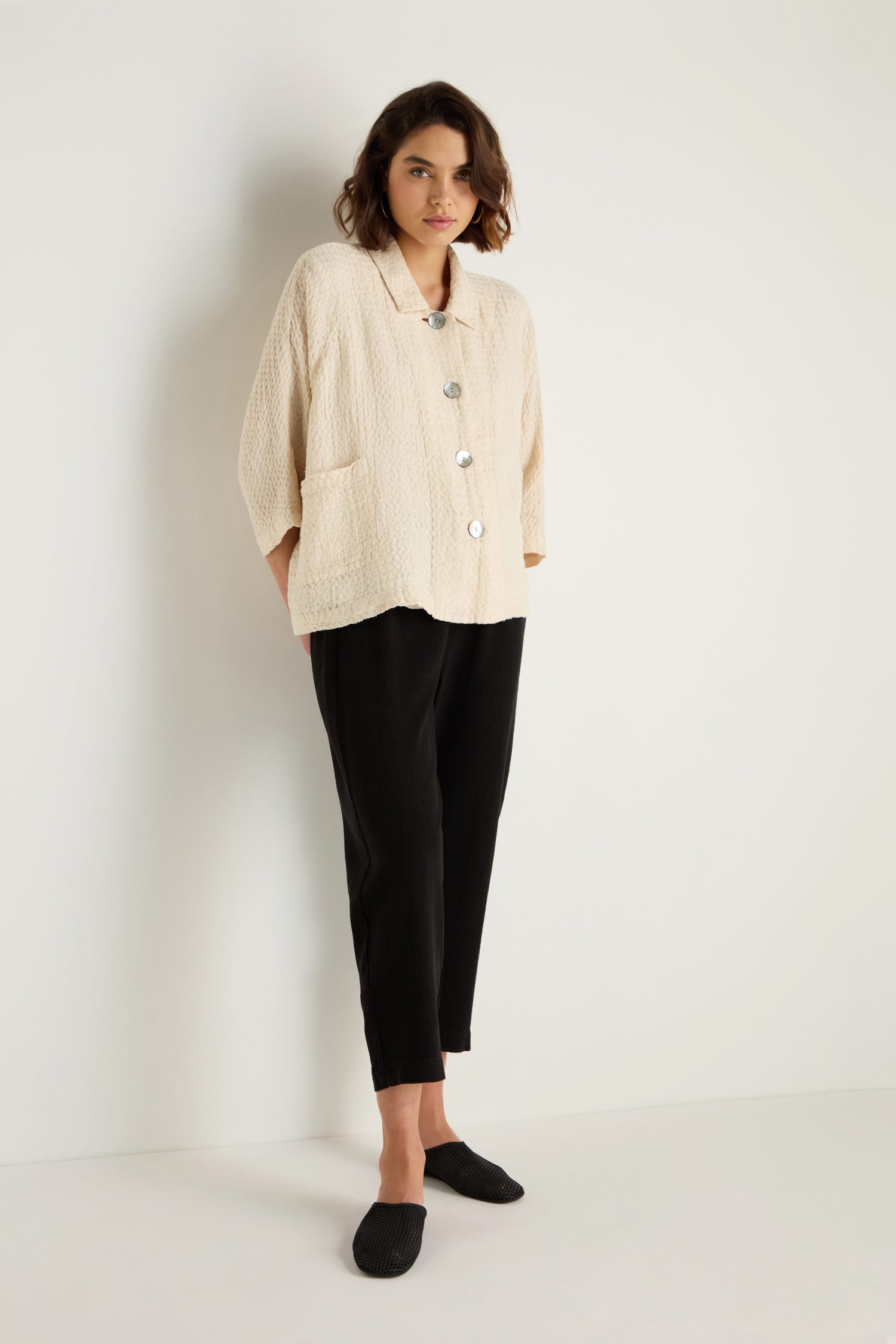 A person stands against a white wall wearing a cream textured button-up jacket, Twisted Linen Slim Trouser in black, and black slip-on shoes.