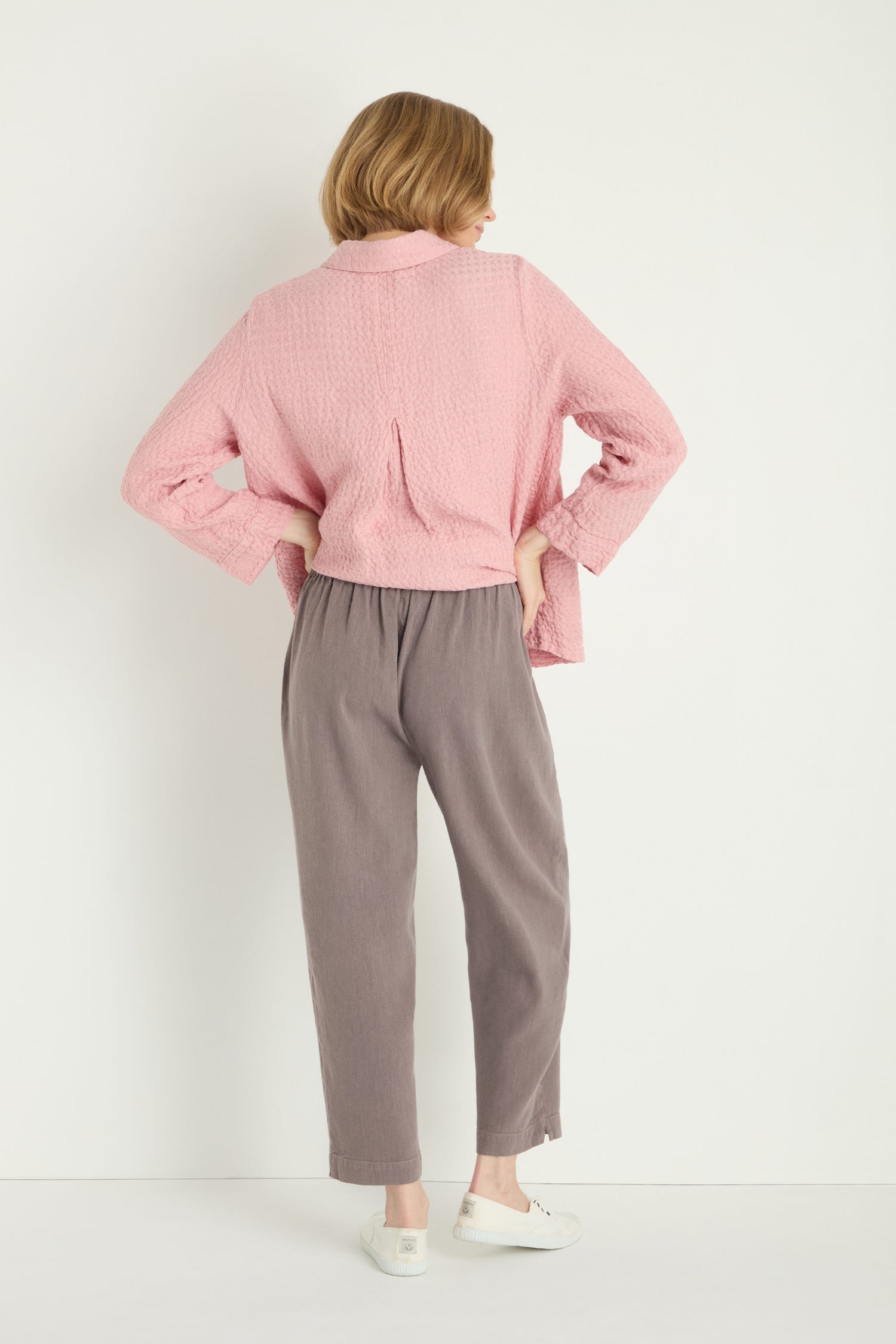 Person stands against a white wall, facing away from the camera, wearing a pink textured top, Twisted Linen Slim Trouser in gray, and white shoes.