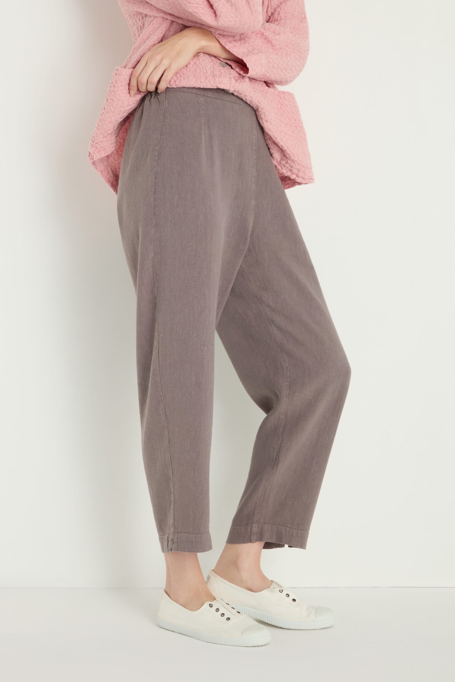 A person stands against a plain light-colored background wearing a pink top, Twisted Linen Slim Trouser in an ankle-grazing length, and white slip-on shoes.