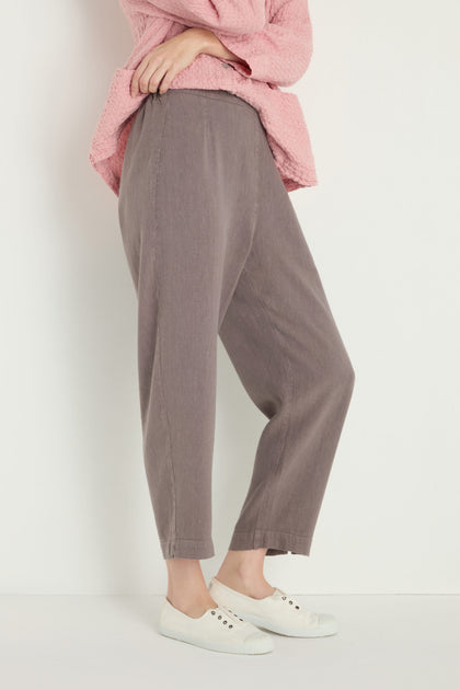 A person stands against a plain light-colored background wearing a pink top, Twisted Linen Slim Trouser in an ankle-grazing length, and white slip-on shoes.