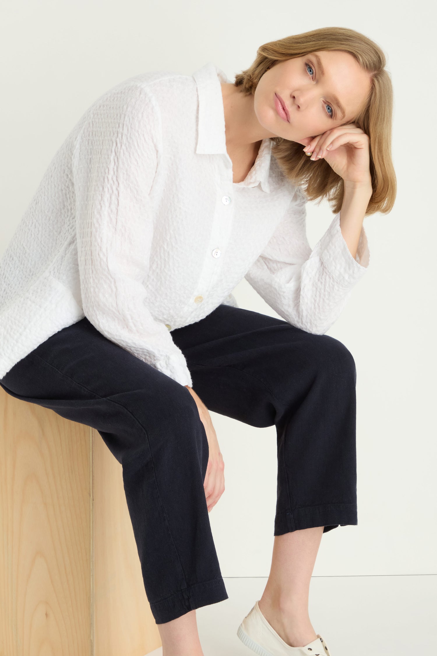 A person with short blonde hair wears a textured white shirt, Twisted Linen Slim Trouser, and white shoes while sitting on a wooden block, resting their head on one hand.