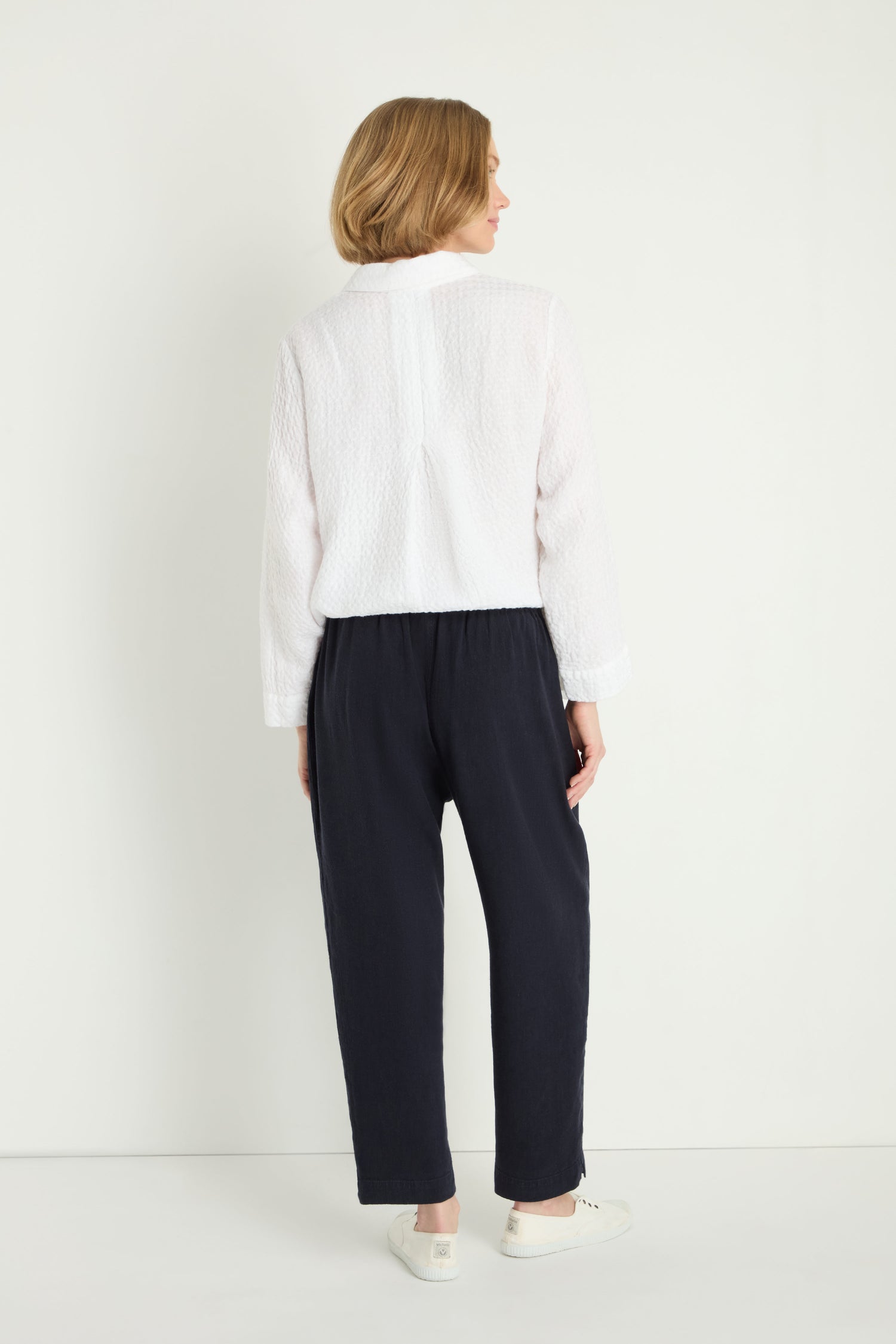 A woman stands facing away wearing a white textured long-sleeve top, Twisted Linen Slim Trouser in navy, and white sneakers against a plain white background.