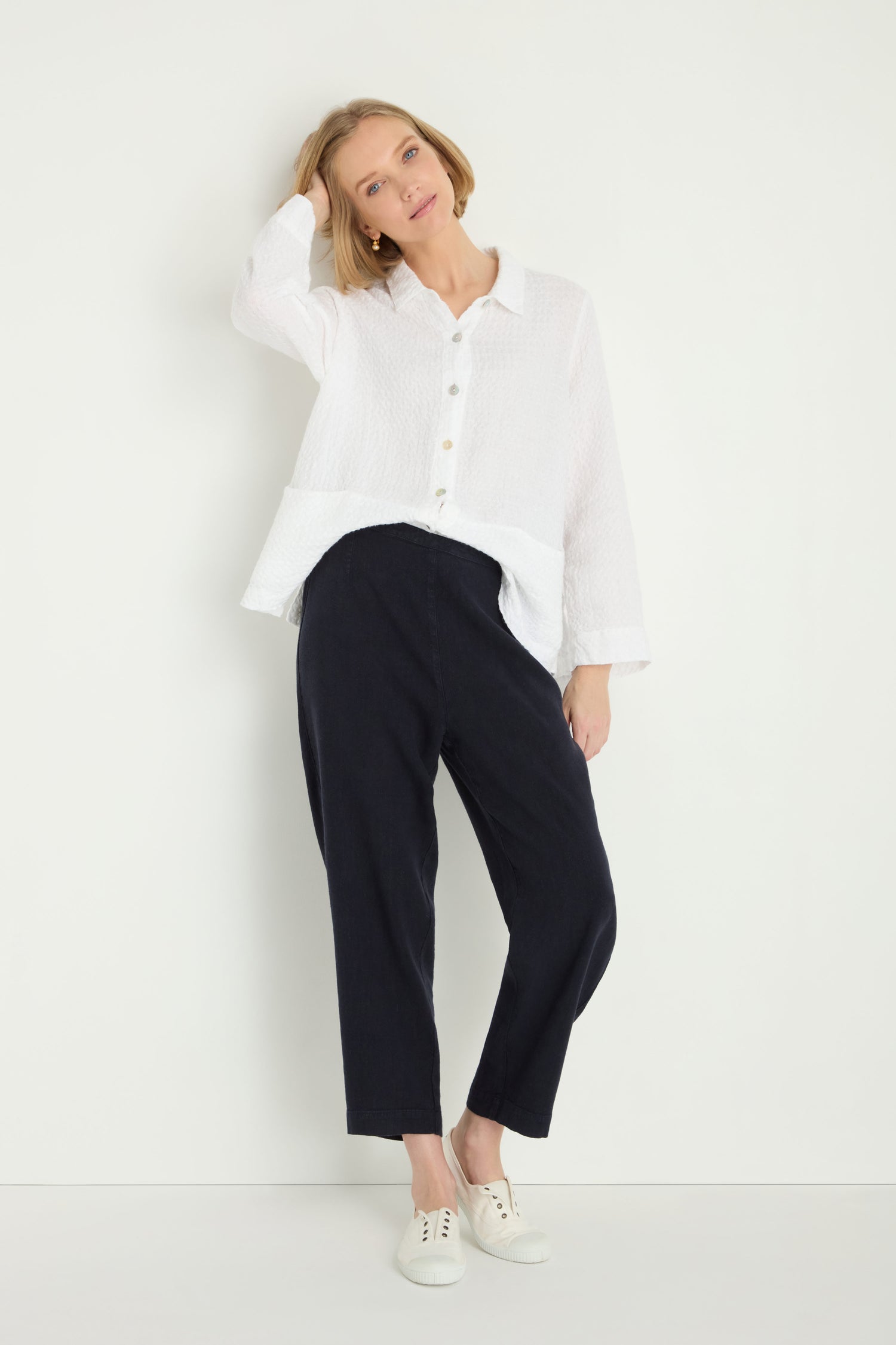 A woman stands against a white wall wearing a loose white button-up shirt, Twisted Linen Slim Trousers, and white sneakers, with one hand touching her head.
