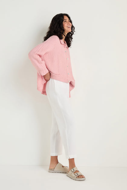Woman standing sideways against a plain wall, wearing a loose pink button-up shirt, Twisted Linen Slim White Trouser at ankle length, and beige sandals.