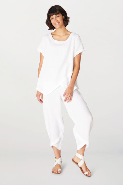 A woman stands smiling in a loose white short-sleeve top and Linen Bubble Trouser_AW23 with an elasticated waistband, paired with white sandals against a plain light background.