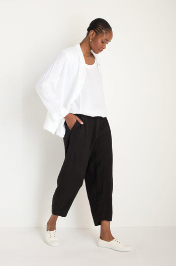 A person stands against a plain background, wearing a white shirt over a white top, Twisted Linen Crop Bubble Trousers for a breathable feel, and white sneakers.