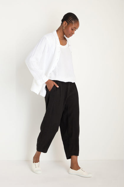 A person stands against a plain background, wearing a white shirt over a white top, Twisted Linen Crop Bubble Trousers for a breathable feel, and white sneakers.