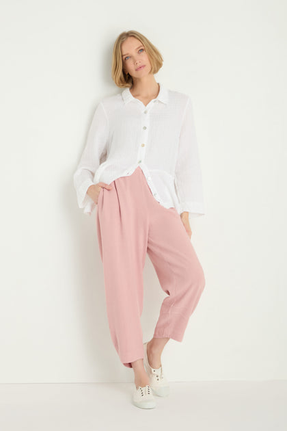 A woman stands against a white wall wearing a white button-up shirt, Twisted Linen Crop Bubble Trouser in loose pink for a breathable feel, and white sneakers, with her hands in her pockets.
