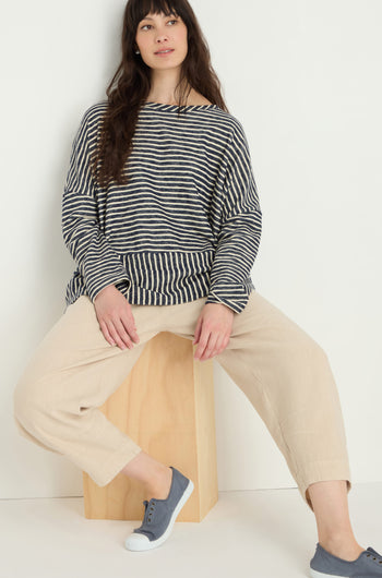 A woman wearing a black and white striped long-sleeve top, Twisted Linen Crop Bubble Trouser in beige, and blue sneakers sits on a wooden block against a plain white background.