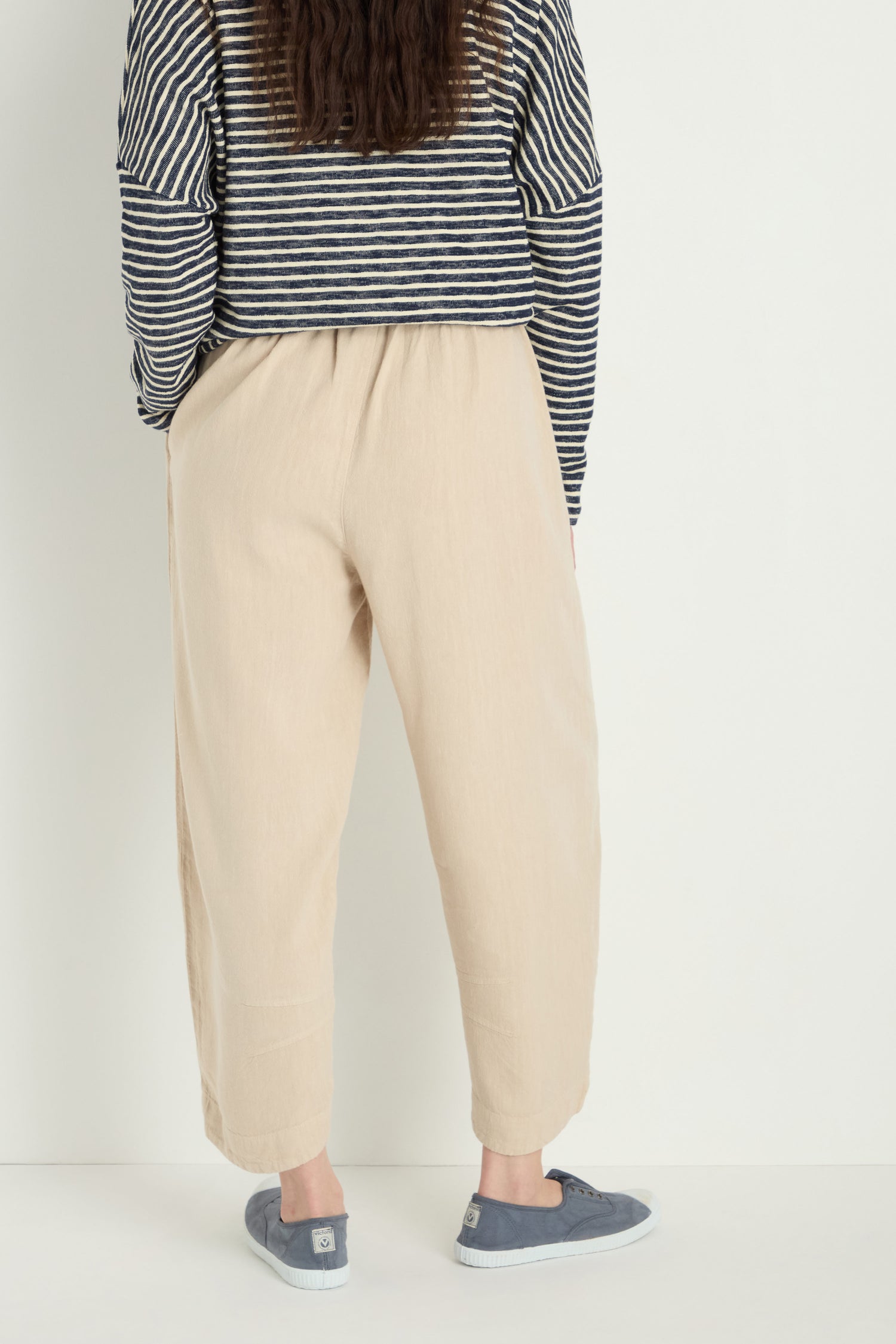 A person is seen from the back against a light background, wearing blue slip-on shoes, a black and white striped top, and Twisted Linen Crop Bubble Trouser in beige with a relaxed bubble silhouette.