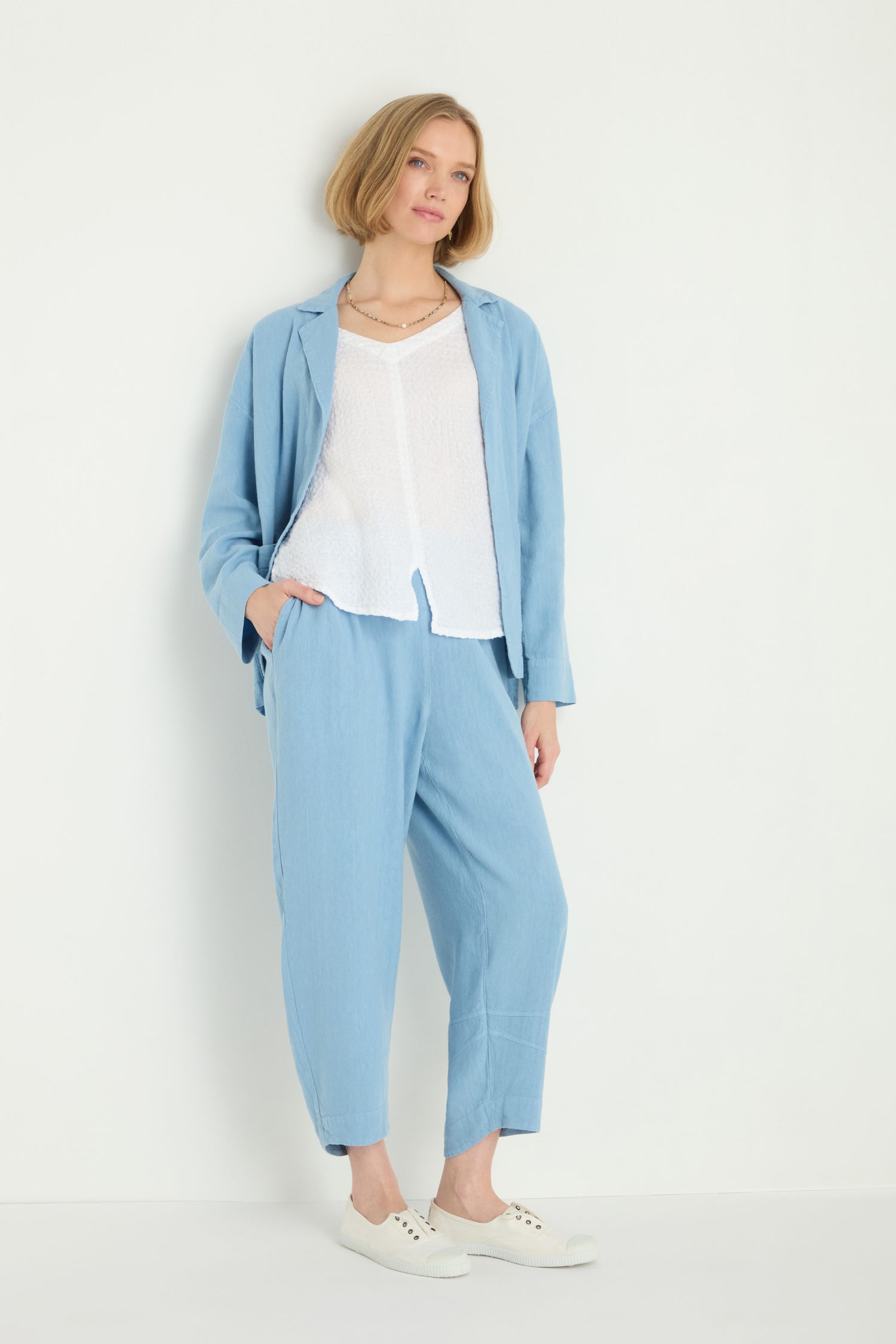 A woman stands against a white wall wearing the Twisted Linen Crop Bubble Trouser in light blue, paired with a white top and white sneakers, with one hand in her pocket.