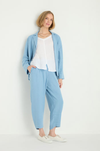 A woman stands against a white wall wearing the Twisted Linen Crop Bubble Trouser in light blue, paired with a white top and white sneakers, with one hand in her pocket.