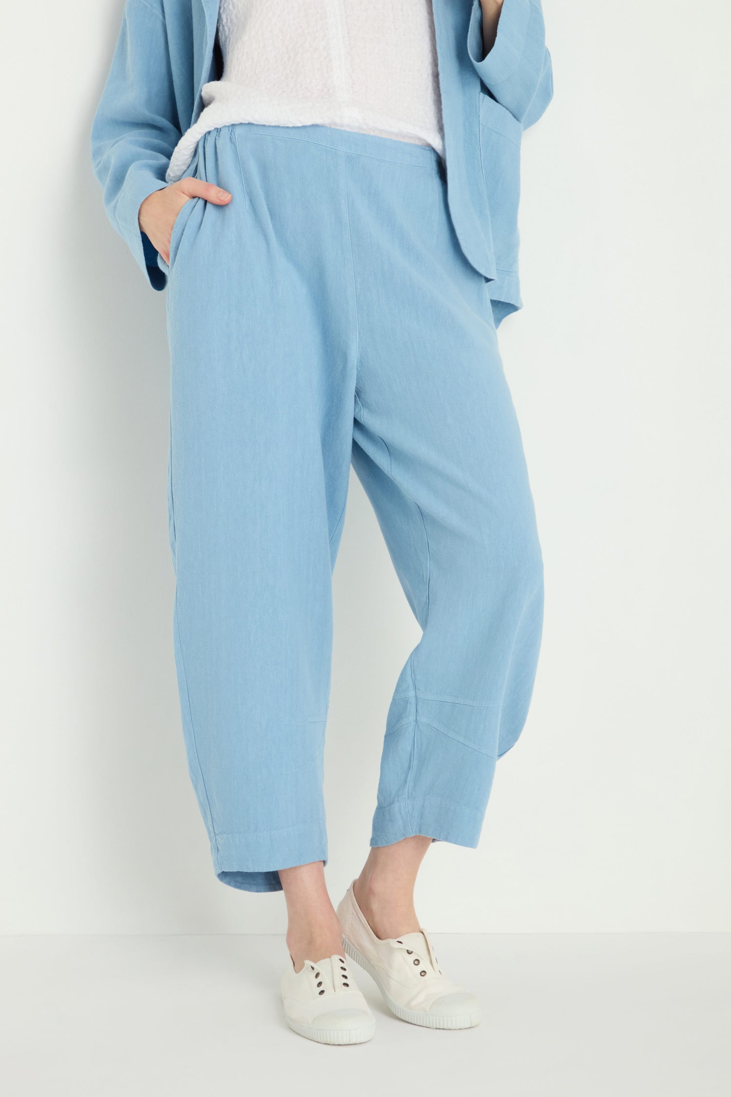 Person wears Twisted Linen Crop Bubble Trouser in light blue with a matching jacket, a white textured top, and white slip-on sneakers, standing against a plain white background.
