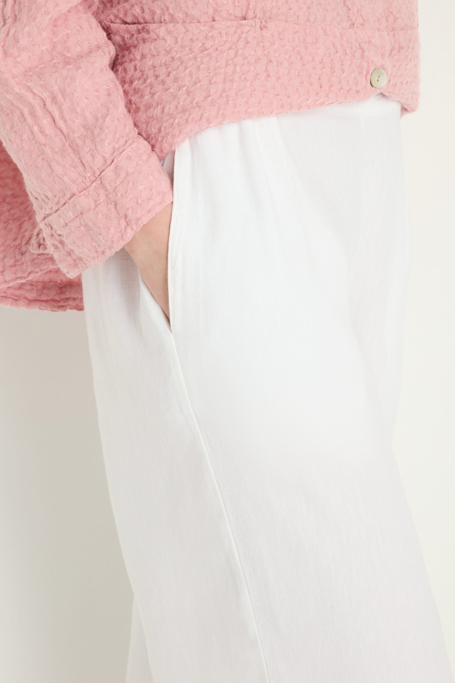 Model stands against a plain background, hand in pocket, wearing white Twisted Linen Crop Bubble Trouser and a pink textured top.