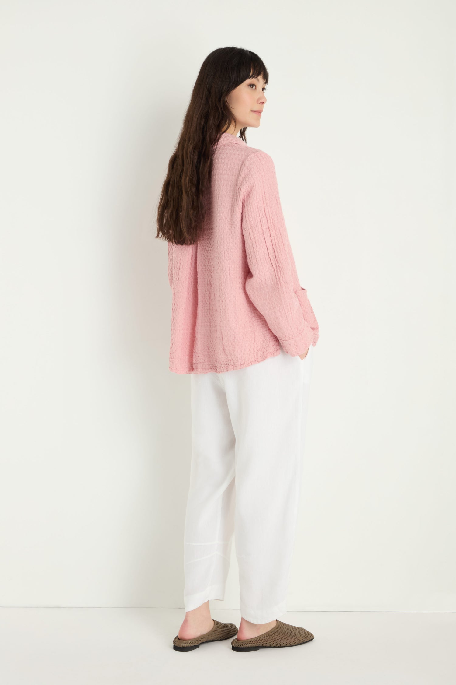 A woman stands sideways against a plain white background, wearing a textured pink sweater, Twisted Linen Crop Bubble Trousers for a breathable feel, and brown slip-on shoes.