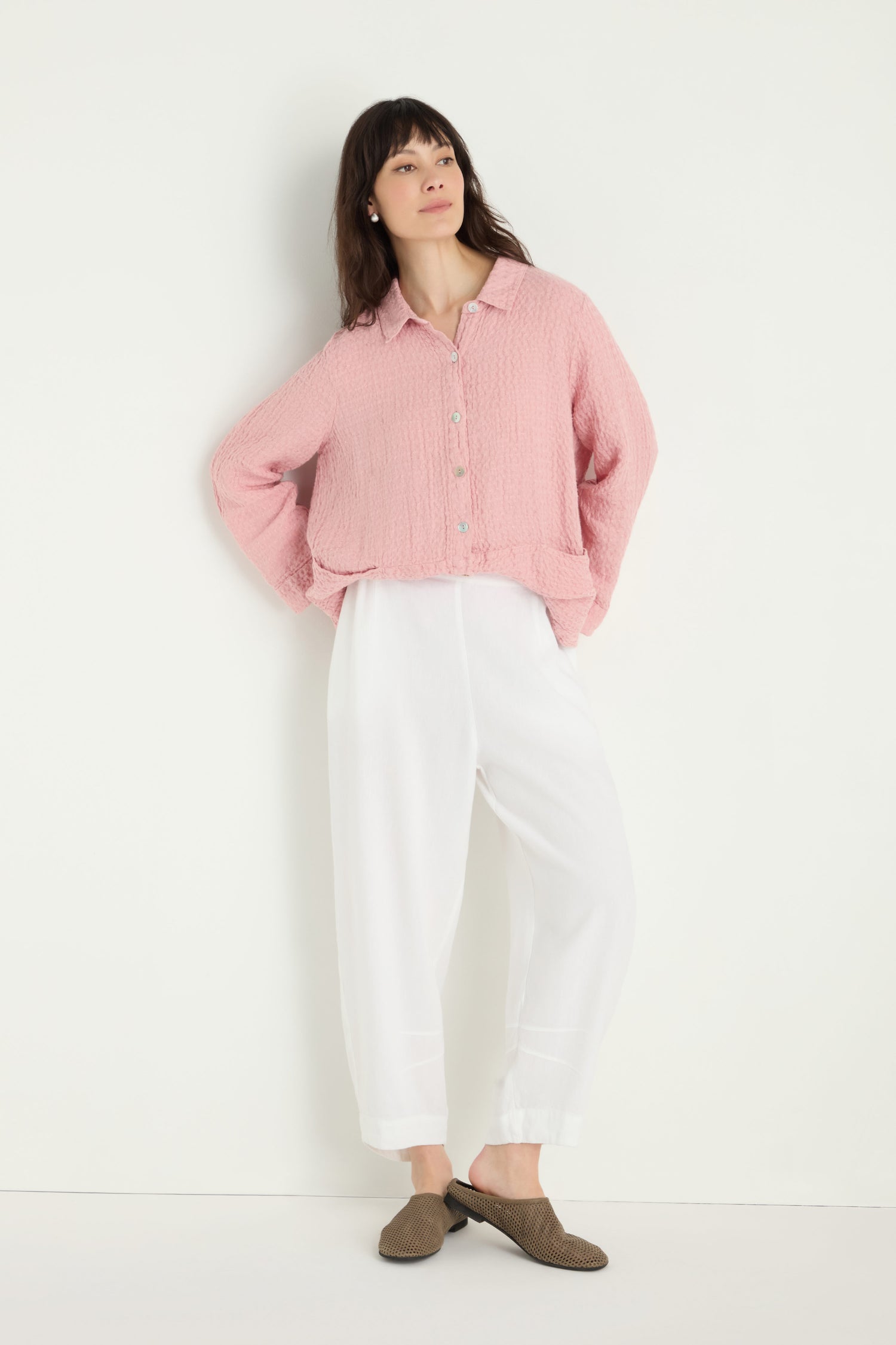A woman stands against a white wall, hands in pockets, wearing a loose pink button-up top, brown slippers, and the Twisted Linen Crop Bubble Trouser in white with a breezy bubble silhouette, gazing slightly upward.
