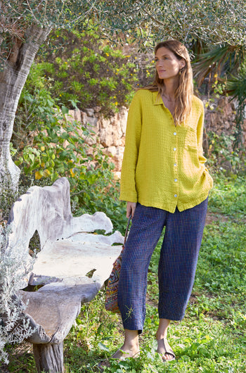 A person in a yellow shirt and Navy Space Dye Check Bubble Trousers stands by a rustic bench in a lush garden.