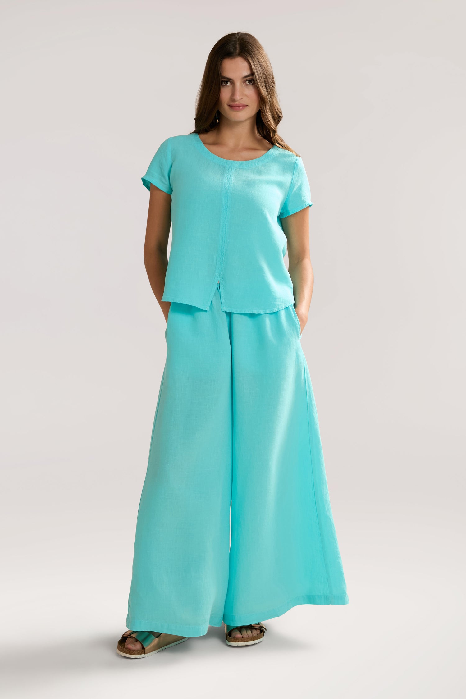 A woman stands wearing a matching turquoise outfit with a short-sleeved top and Organza Linen Wide Trouser. She has her hands in her pockets and is wearing sandals, exuding an air of relaxed luxury.