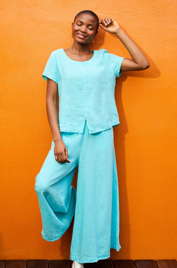 A person in a loose-fitting, light blue outfit with Organza Linen Wide Trouser stands against an orange background, smiling and raising one hand.