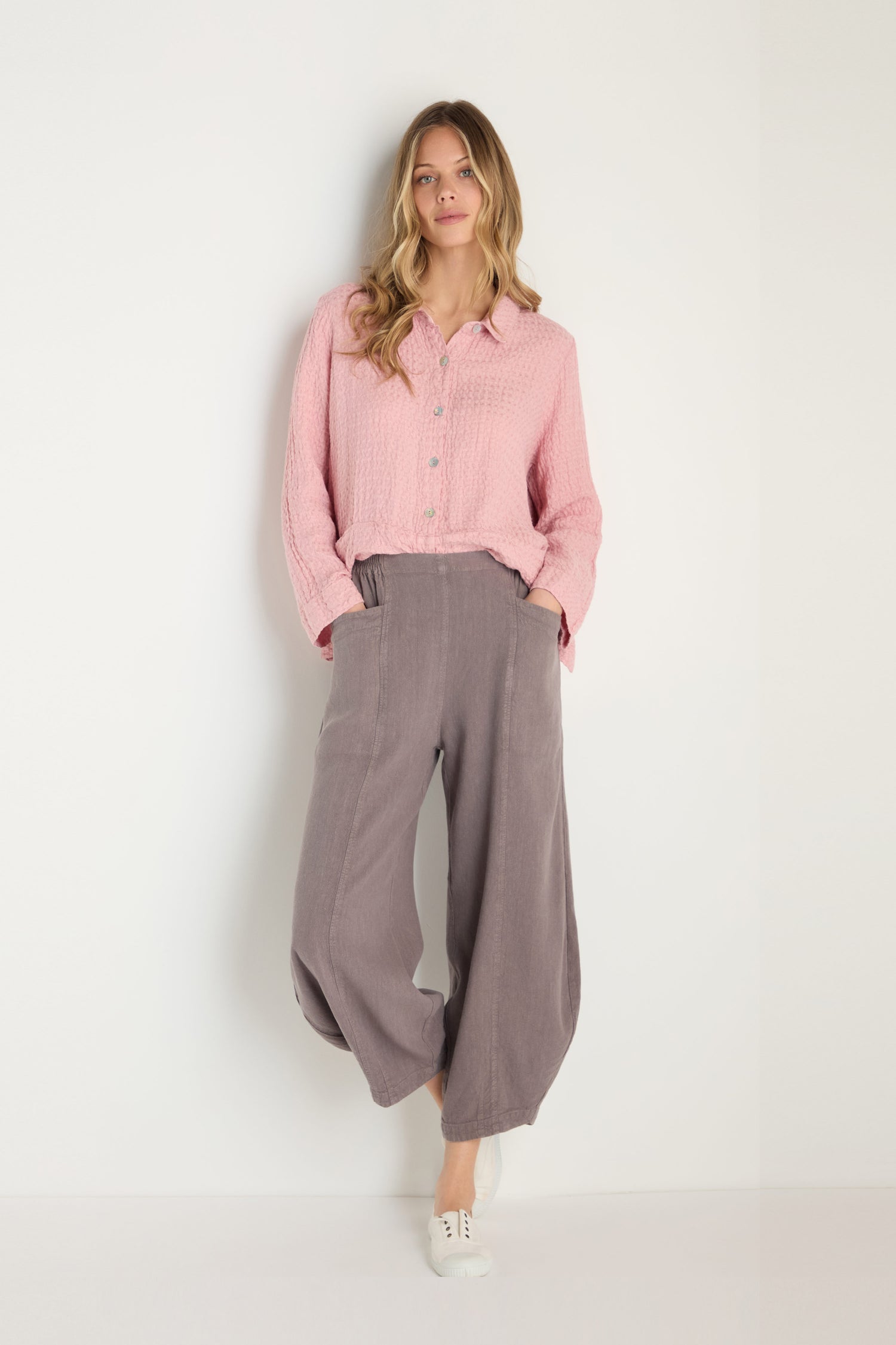 A woman stands against a white wall wearing a pink button-up shirt, Twisted Linen Bubble Trouser in gray, and white shoes, with her hands in her pockets.