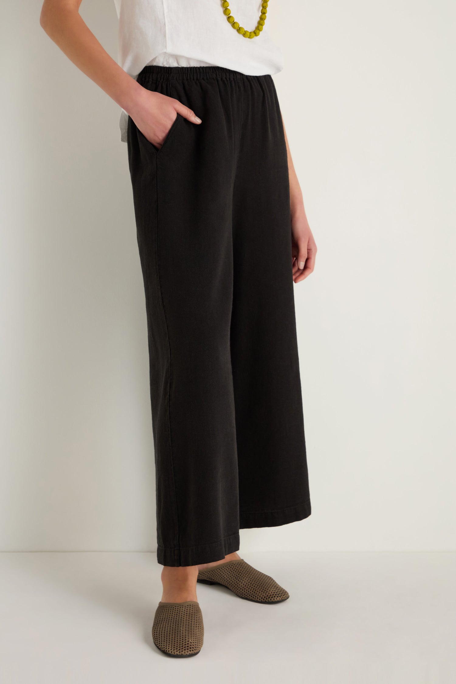 A person wearing black Twisted Linen Wide Trouser, a white shirt, a yellow beaded necklace, and brown textured slip-on shoes stands against a plain light background.