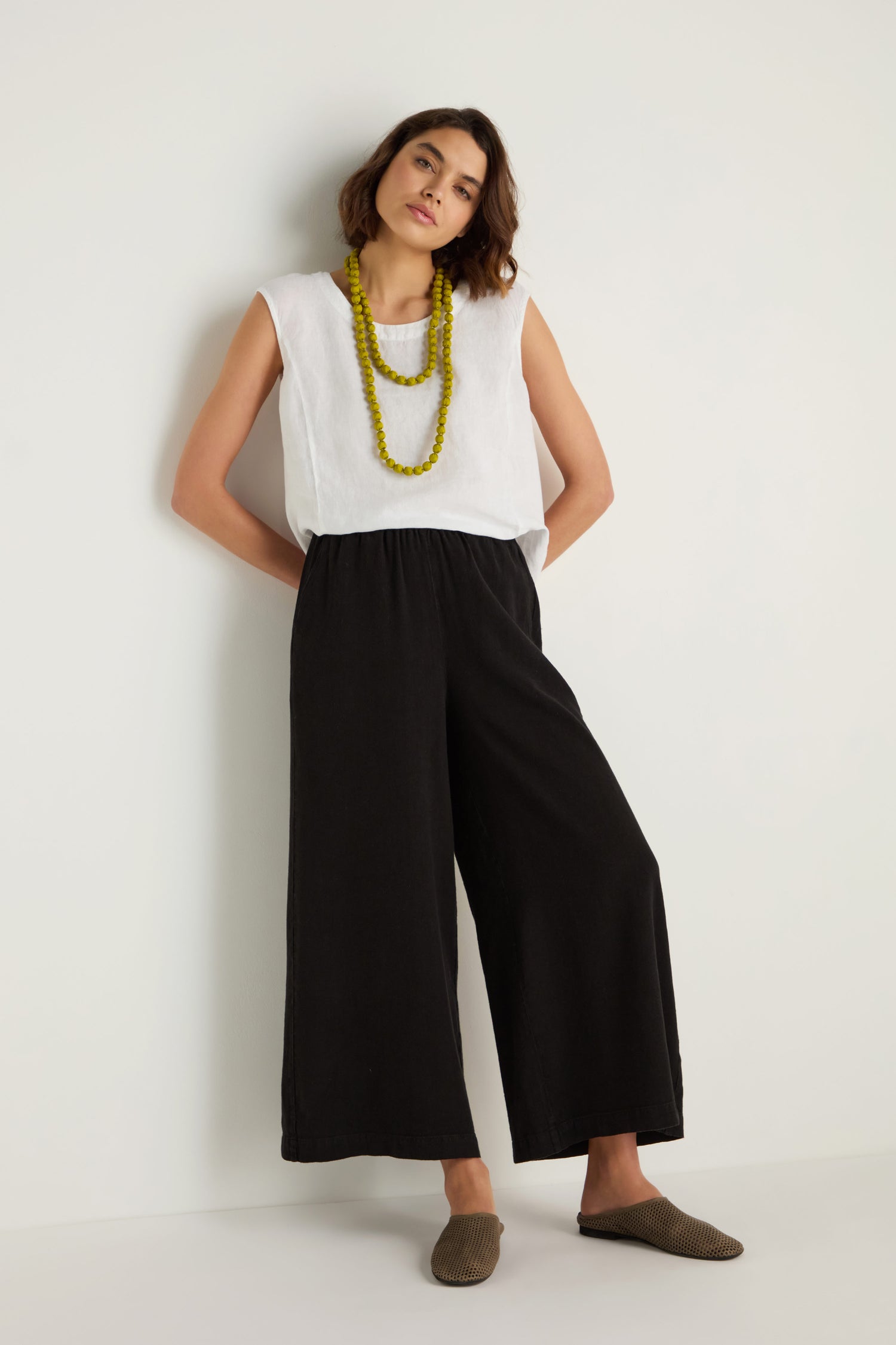 A person stands against a white wall wearing a white sleeveless top, Twisted Linen Wide Trouser in black, brown slip-on shoes, and a long yellow beaded necklace.