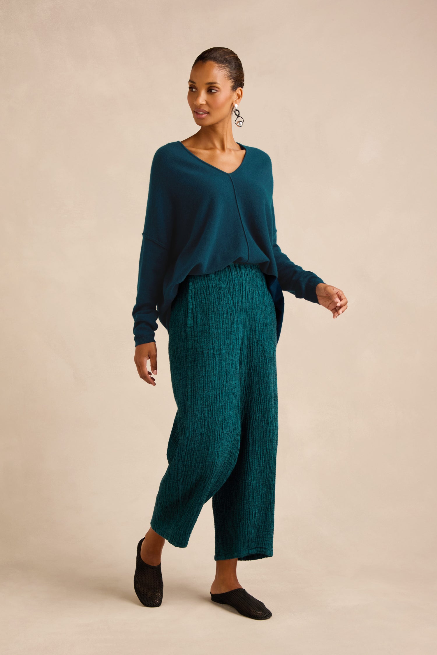 Woman wearing a teal V-neck sweater, Crinkle Linen Blend Bubble Trousers in textured teal, and black slip-on shoes stands against a beige background.