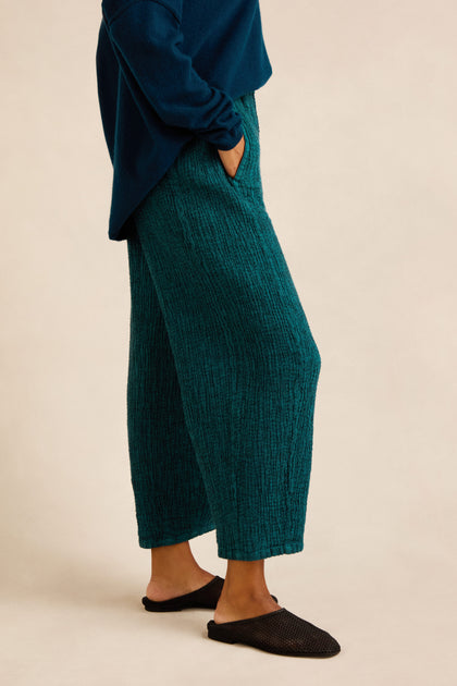 A person wearing Crinkle Linen Blend Bubble Trousers in textured teal, paired with a matching long-sleeve top and black slip-on shoes, stands with one hand in their pocket against a neutral background.
