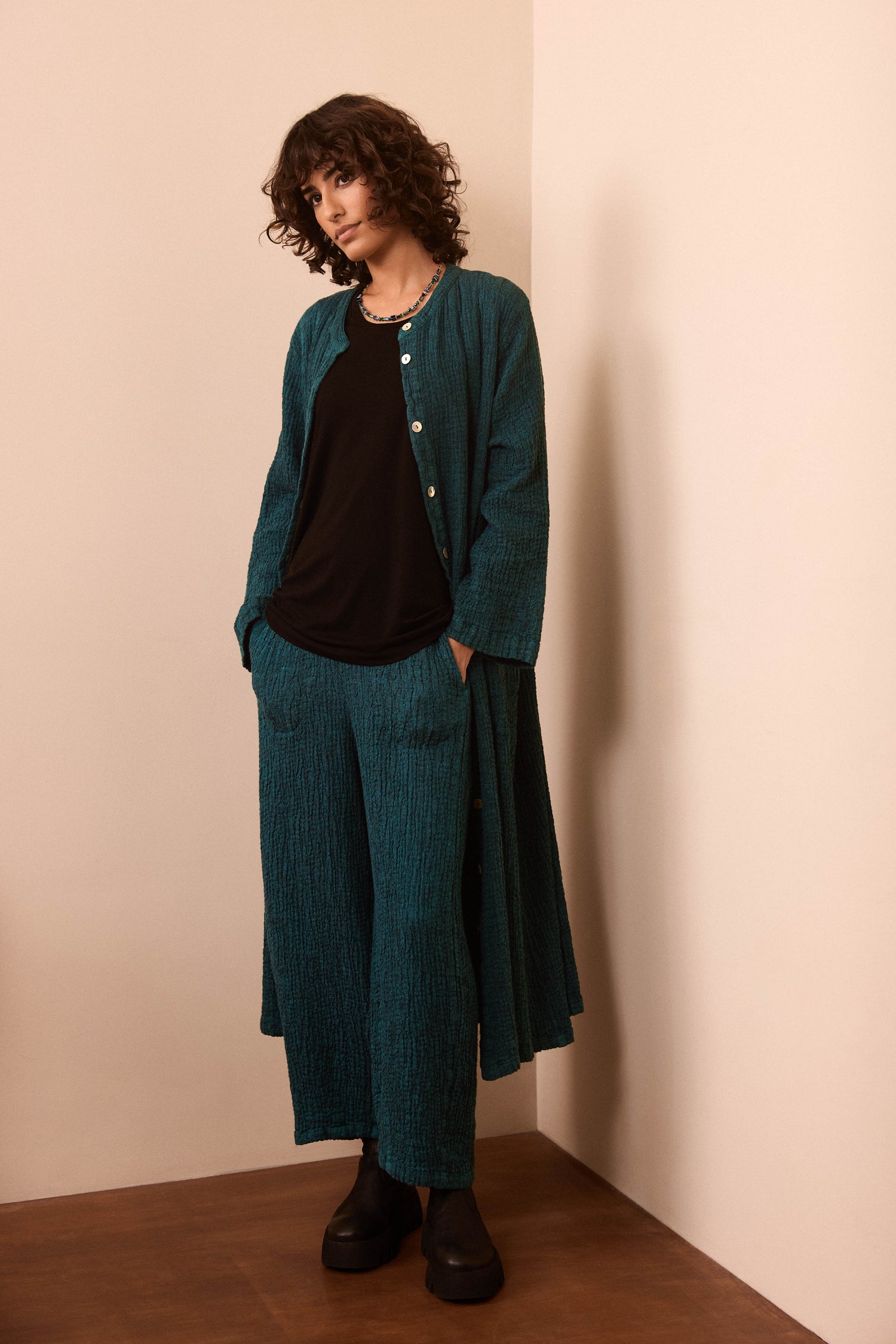 Person wearing a teal textured fabric long jacket and matching Crinkle Linen Blend Bubble Trousers with an elasticated waist over a black shirt, standing indoors against a beige wall, hands in pockets and maintaining a neutral expression.