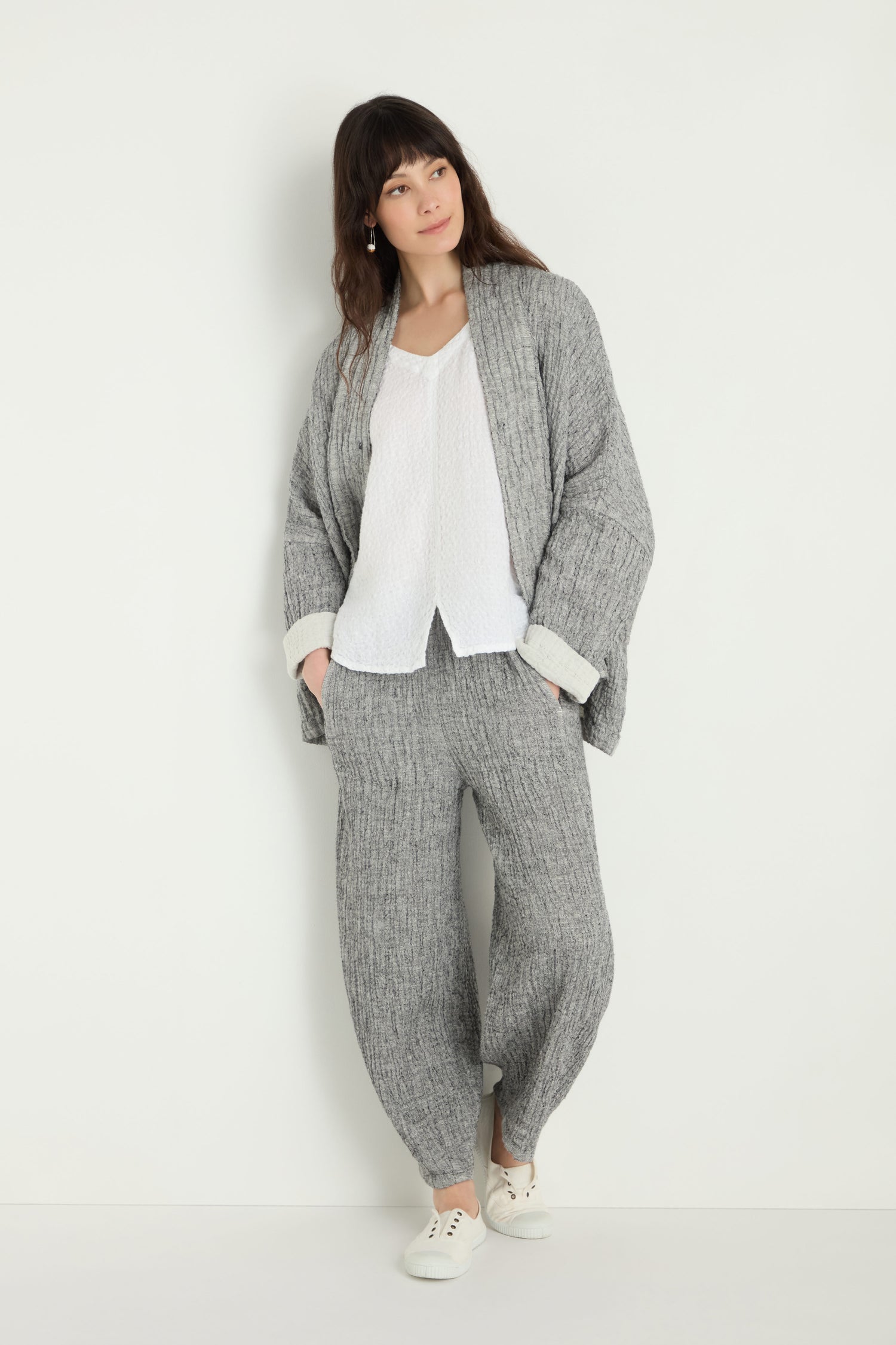 A woman stands against a white wall wearing a loose grey knit cardigan, Crinkle Double Face Linen Bubble Trouser, a white blouse, and white sneakers.