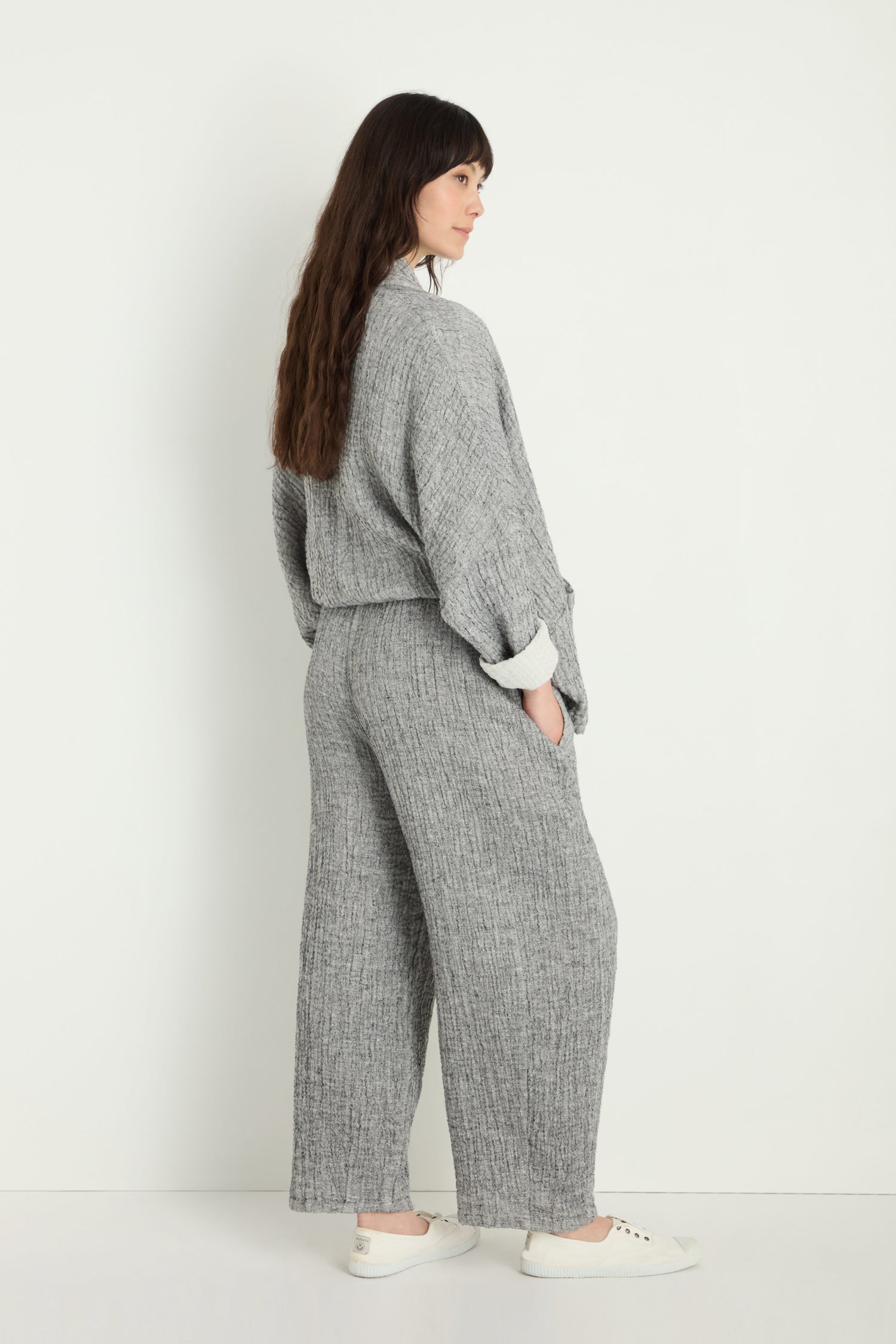 Person stands against a white wall, wearing Crinkle Double Face Linen Bubble Trouser in textured grey with white sneakers, hands in pockets and facing slightly away from the camera.