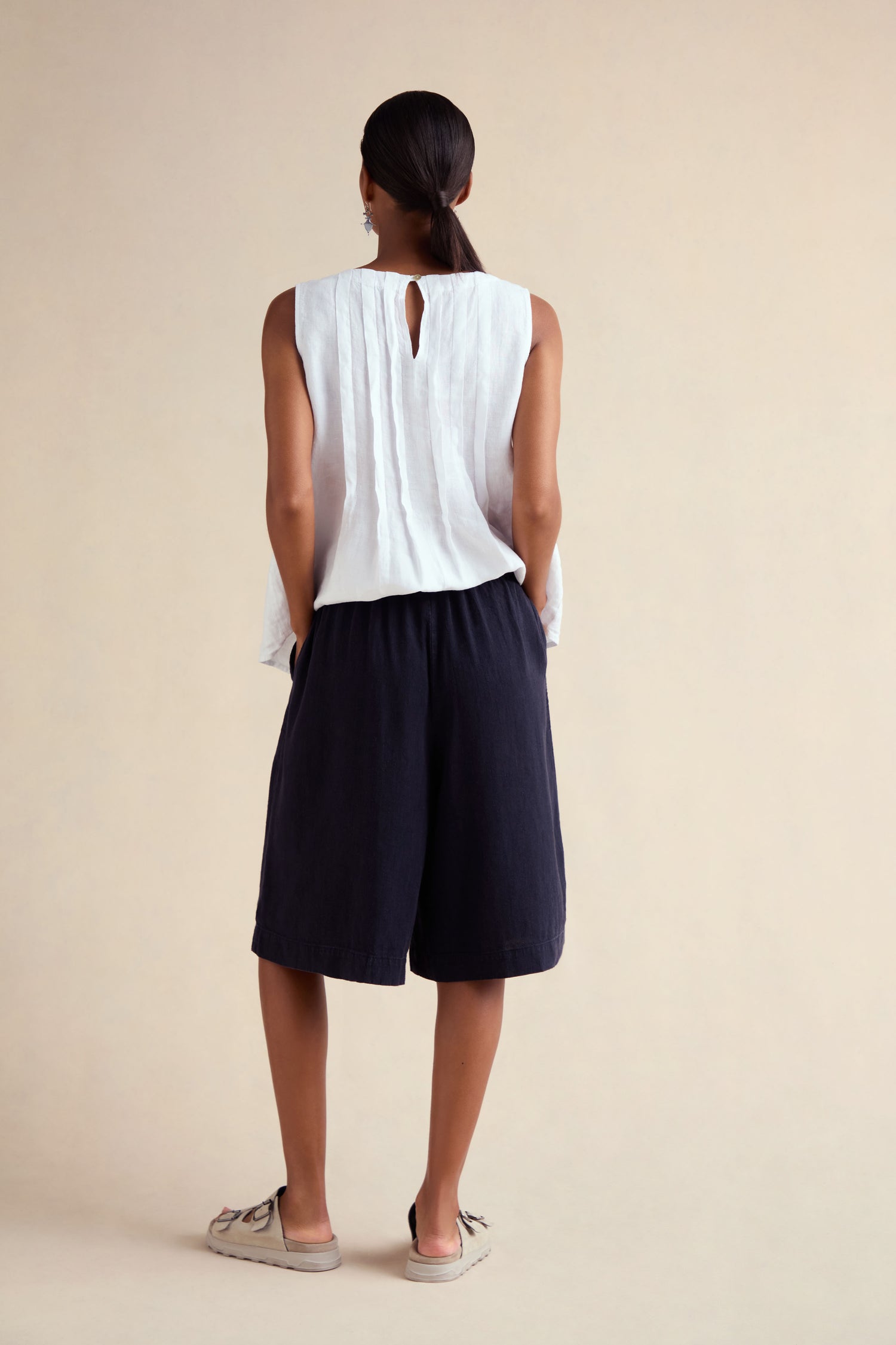 A woman stands with her back to the camera, wearing a sleeveless white top and Twisted Linen Wide Shorts, paired with beige flats, against a plain light background.