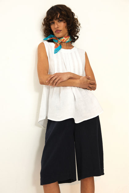 A woman stands against a white wall with crossed arms, wearing the Twisted Linen Wide Shorts featuring an elasticated waist, paired with a sleeveless white top and a colorful scarf around her neck.