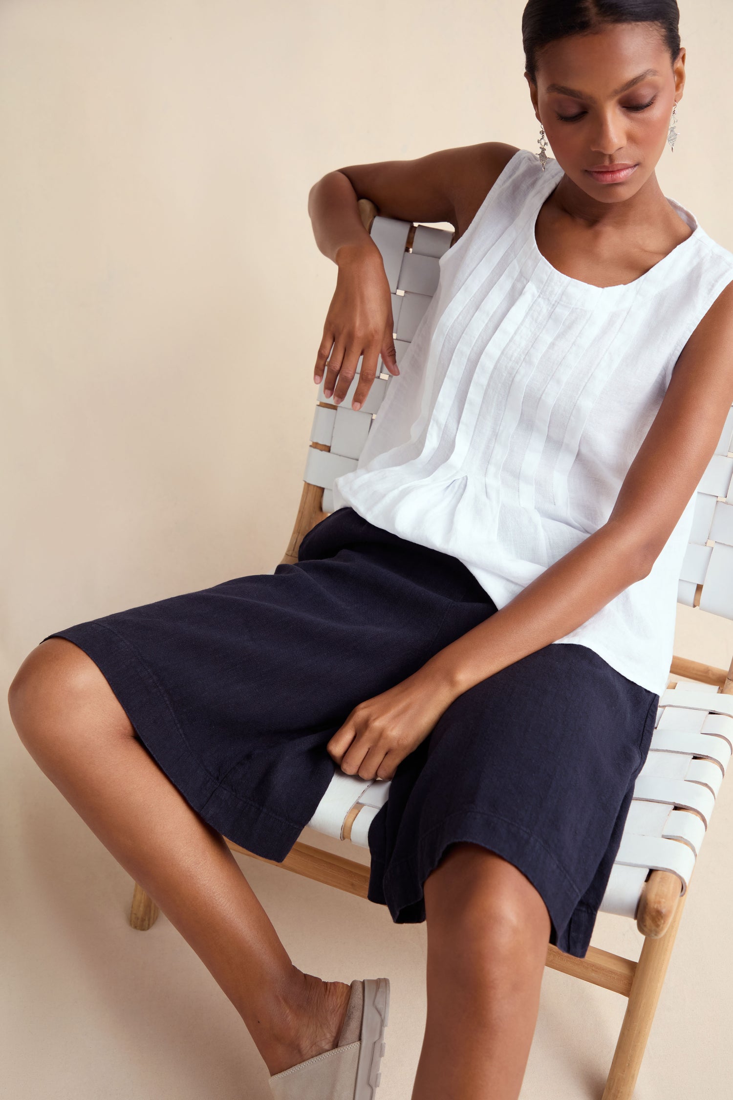 A woman sits on a woven chair wearing Twisted Linen Wide Shorts with a sleeveless white blouse and light slip-on shoes, set against a plain background.