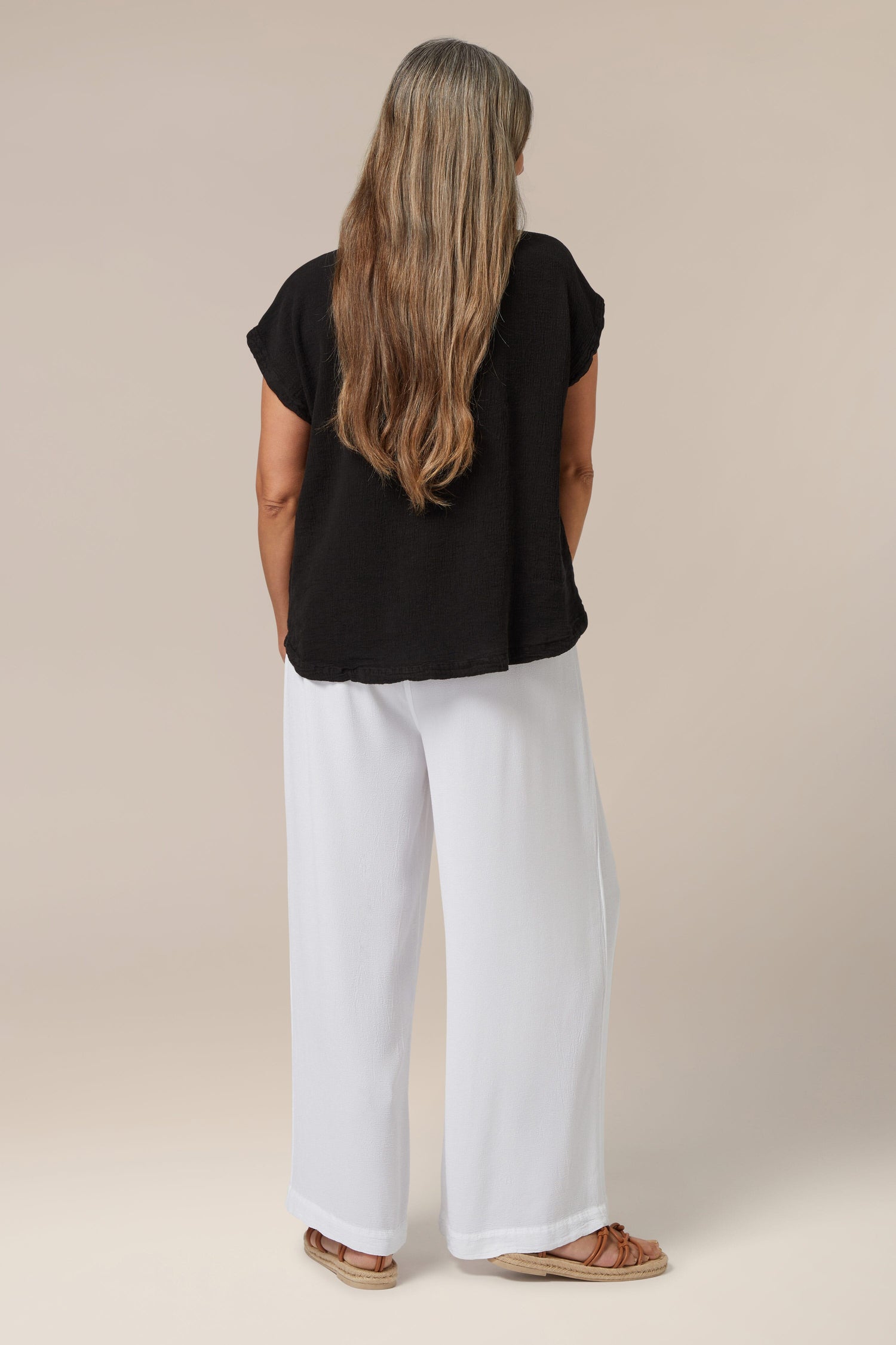 Sentence with product name: Woman standing with her back to the camera, wearing a Sunny Day Crinkle Linen Top and white trousers, with long brown hair reaching half her back.