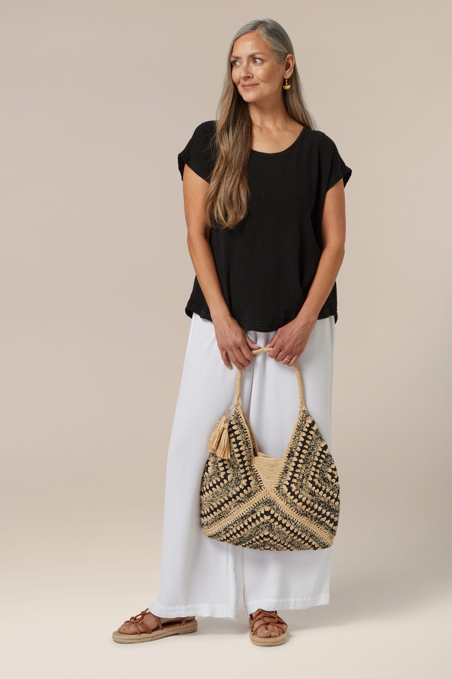Woman standing in a studio wearing a Summer Breeze Top, white pants, holding a woven handbag, and wearing brown sandals for a warm weather look.