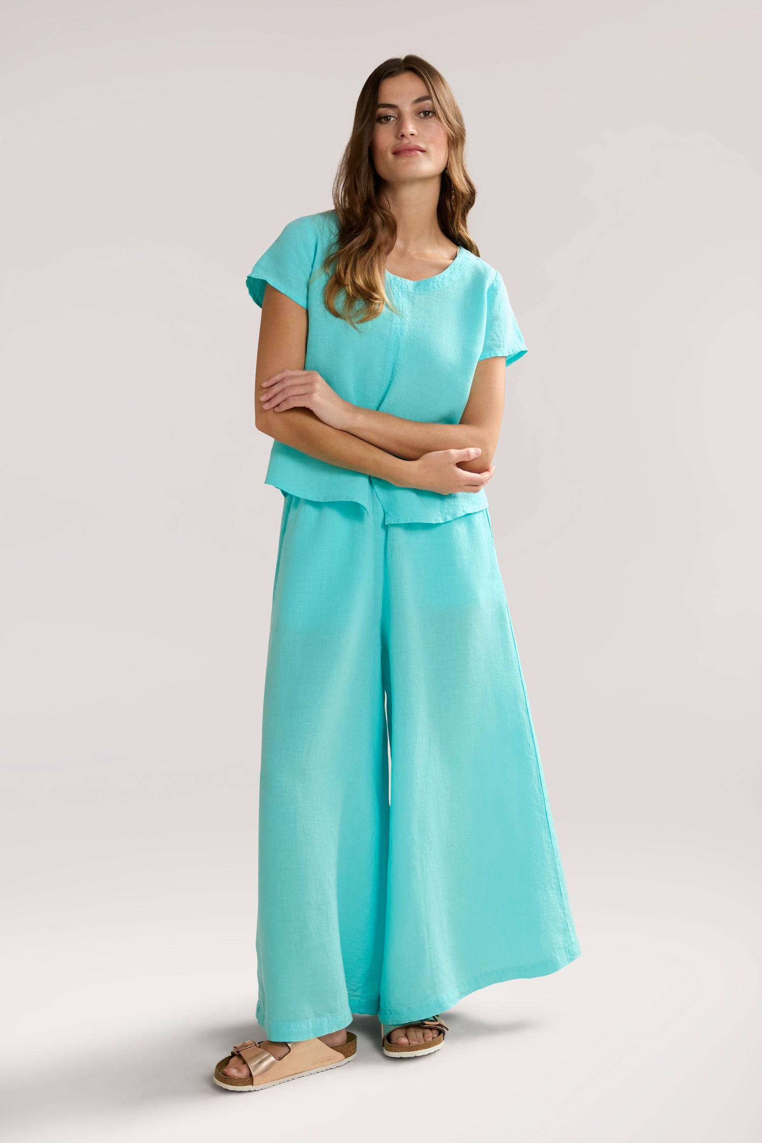 A person stands confidently in a light turquoise outfit consisting of a Slit Hem Linen Top with flutter sleeves and wide-leg pants, paired with light-colored sandals. The background is plain and light, enhancing the outfit's airy silhouette.