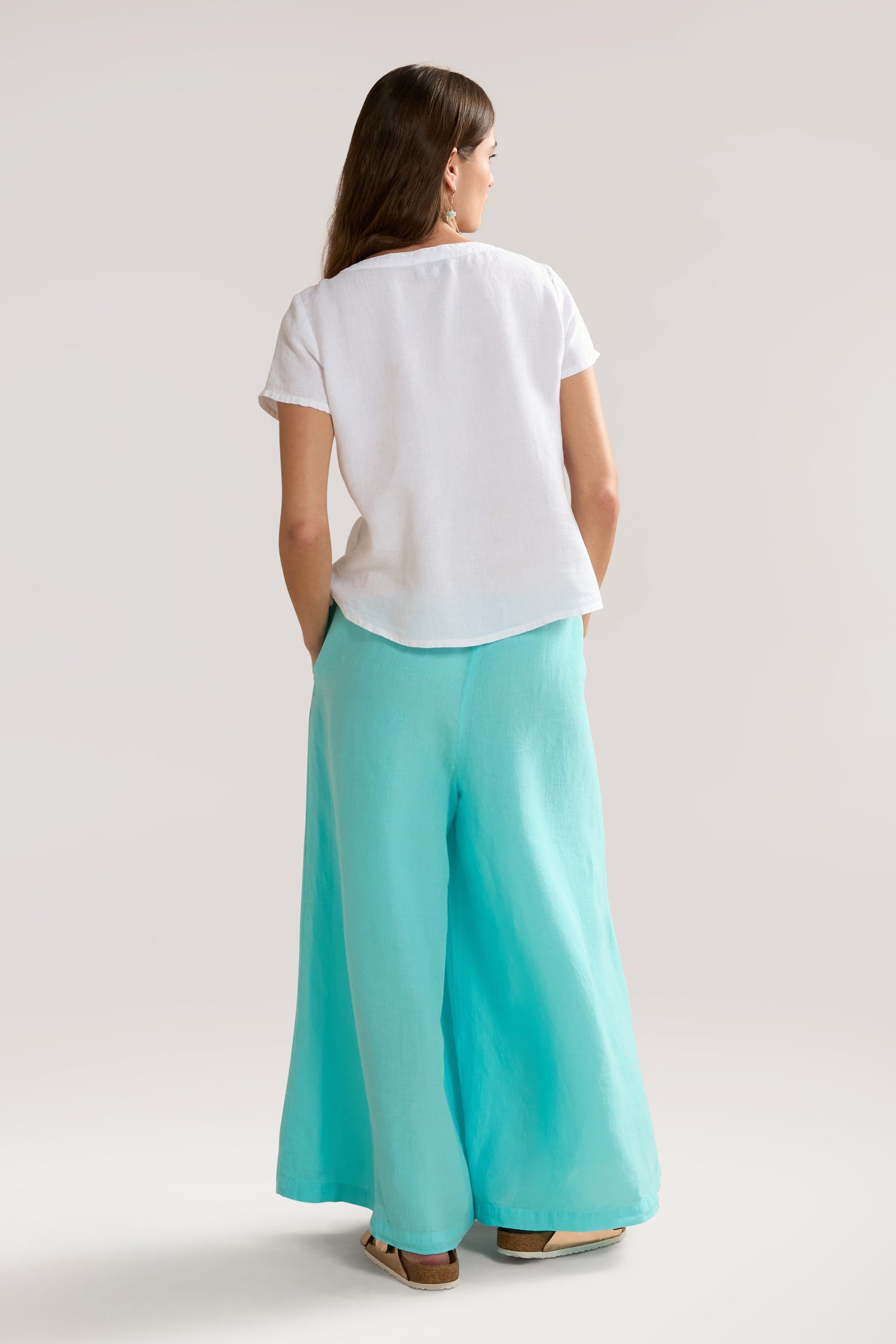 A woman stands with her back to the camera, wearing a Slit Hem Linen Top and turquoise wide-leg pants. Her hands are in her pockets, emphasizing the airy silhouette of her outfit.