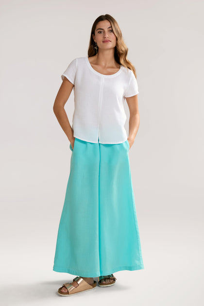 A person stands wearing a white Slit Hem Linen Top with flutter sleeves, turquoise wide-leg pants, and brown sandals, with hands in pockets, against a plain background.